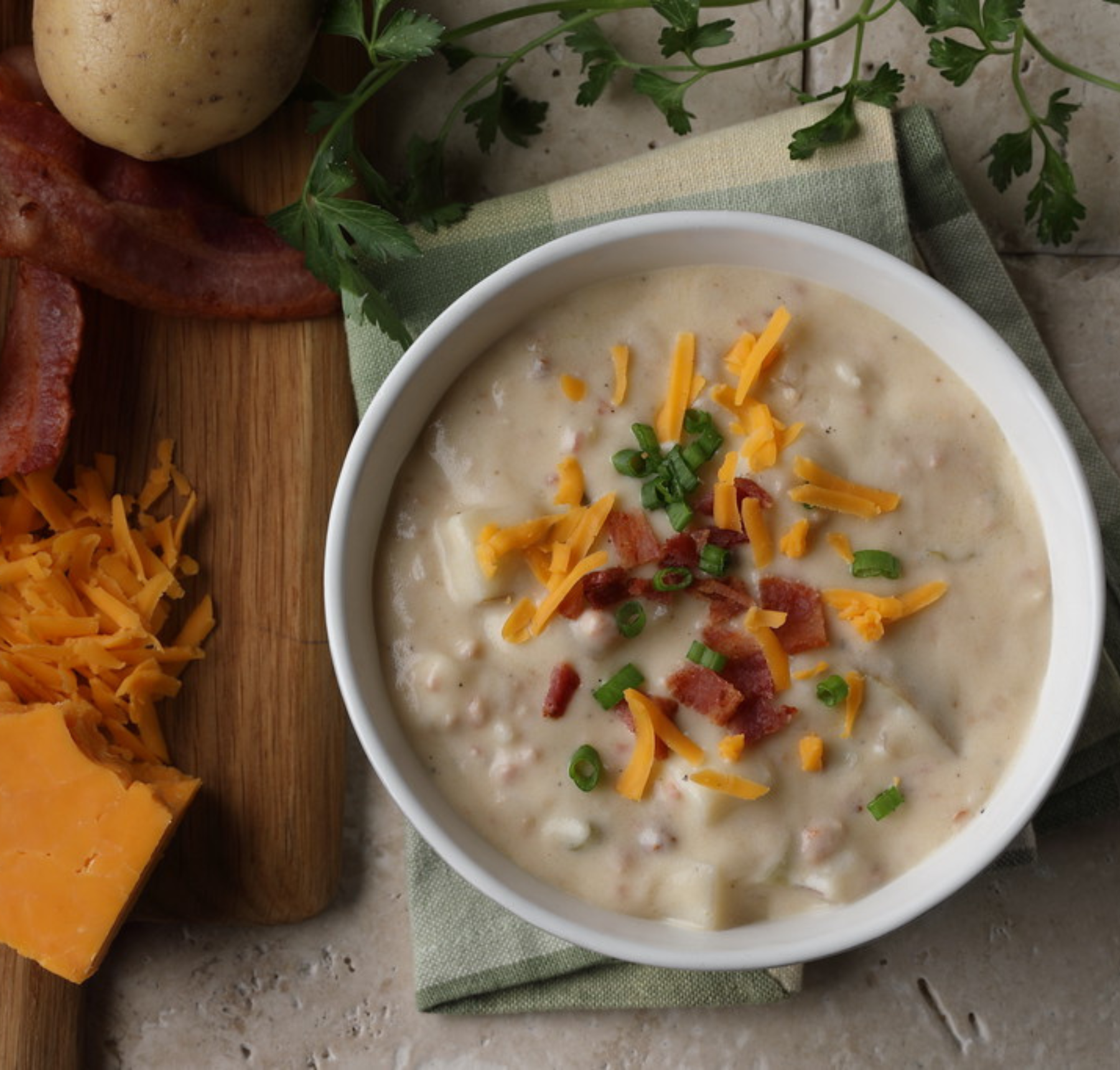 A bowl of baked potato soup garnished with bacon, cheese, and green onions, with ingredients and a napkin in the background.