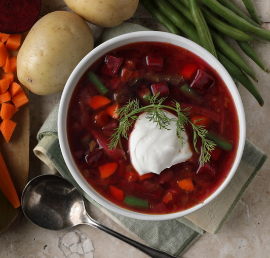 A bowl of vegetable borscht soup topped with sour cream and dill, surrounded by ingredients like beets, carrots, potatoes, and green beans.