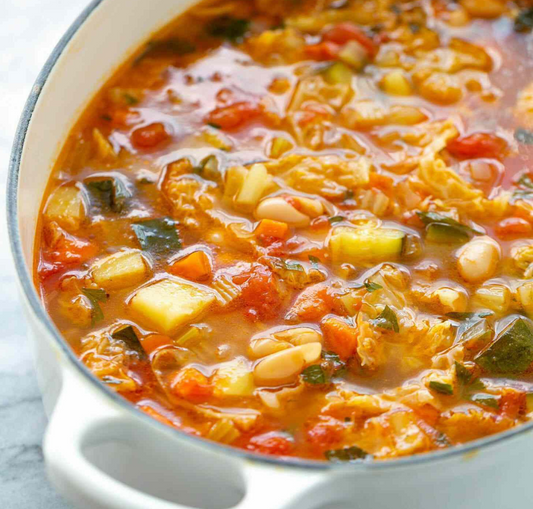 A bowl of vegetable minestrone soup, featuring a variety of vegetables such as spinach, carrots, onions, celery, garlic, green beans, zucchini, and tomatoes, as well as kidney beans, all cooked and served in a white bowl.